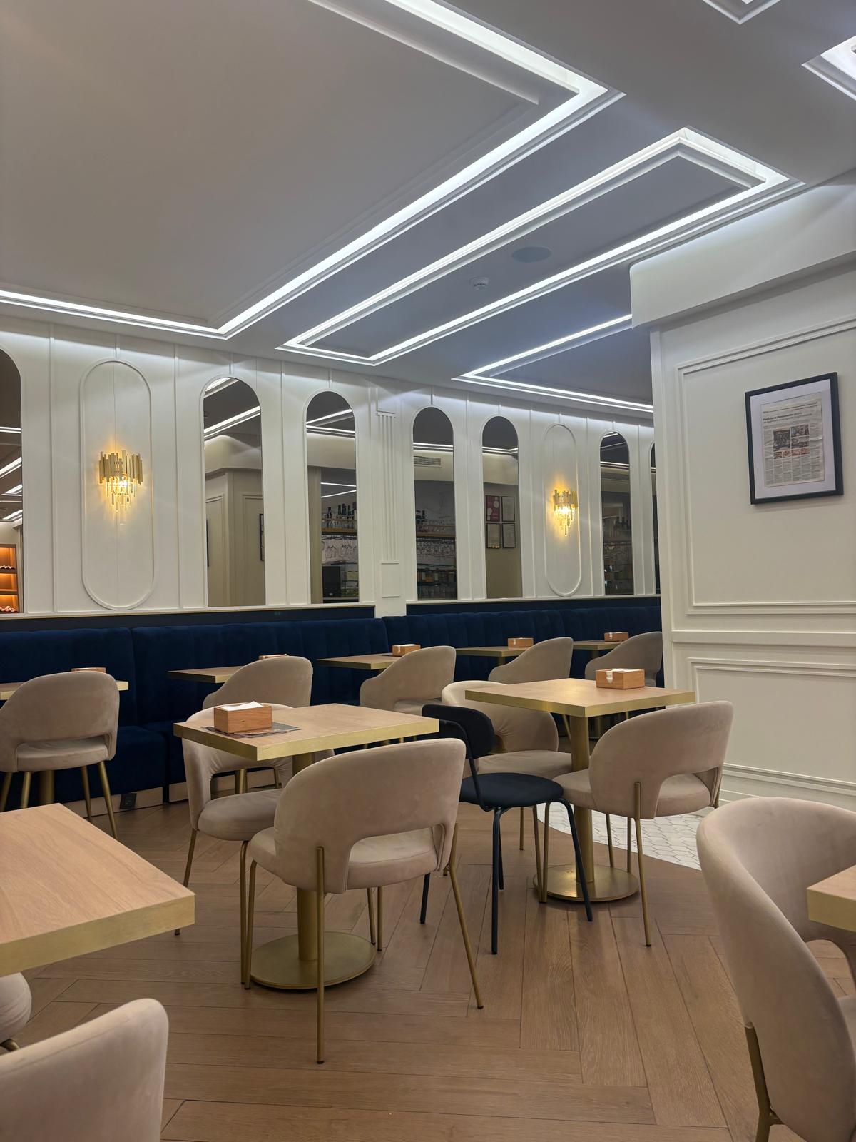 Sala da pastelaria, com mesas de madeira, cadeiras bege e um sofá azul-marinho